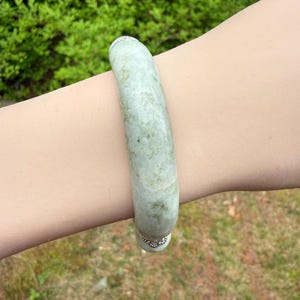 K&ouml;nnte beinhalten: Ein hellgr&uuml;nes Jade-Armreif-Armband. Das Armband ist ein massives, glattes Band mit leicht gesprenkeltem Aussehen. Der Jade hat eine durchscheinende Qualit&auml;t mit subtilen Farbvariationen. Das Armband wird am Handgelenk getragen, mit einem verschwommenen gr&uuml;nen Hintergrund.
