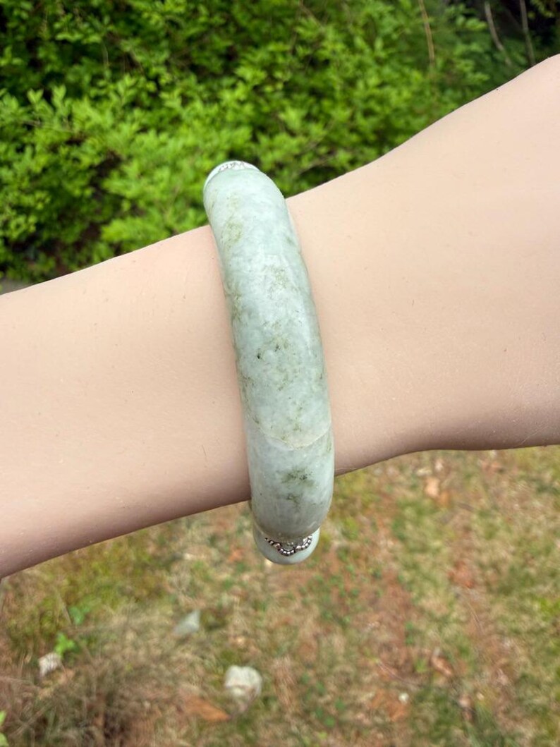 K&ouml;nnte beinhalten: Ein hellgr&uuml;nes Jade-Armreif-Armband. Das Armband ist ein massives, glattes Band mit leicht gesprenkeltem Aussehen. Der Jade hat eine durchscheinende Qualit&auml;t mit subtilen Farbvariationen. Das Armband wird am Handgelenk getragen, mit einem verschwommenen gr&uuml;nen Hintergrund.