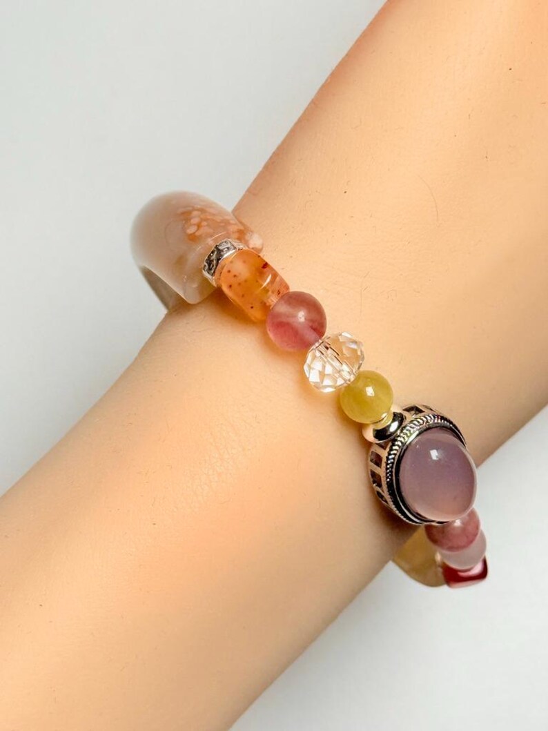 Pu&ograve; includere: Un braccialetto di perline con una variet&agrave; di pietre preziose colorate. Il braccialetto include una grande pietra di colore chiaro, perline arancioni, sfere rosa e gialle e una perla trasparente sfaccettata. Il braccialetto &egrave; indossato su un polso.