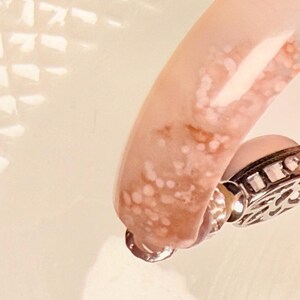 Pu&ograve; includere: Primo piano di un braccialetto rosa e bianco con una chiusura decorativa argentata. Il braccialetto ha un aspetto marmorizzato con un mix di tonalit&agrave; rosa e bianche. La chiusura &egrave; dettagliata con un design ornato.