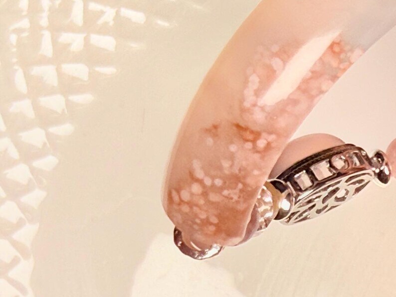 Pu&ograve; includere: Primo piano di un braccialetto rosa e bianco con una chiusura decorativa argentata. Il braccialetto ha un aspetto marmorizzato con un mix di tonalit&agrave; rosa e bianche. La chiusura &egrave; dettagliata con un design ornato.