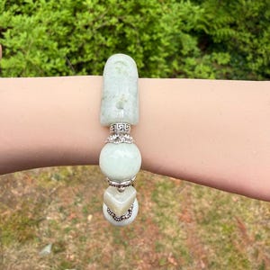 K&ouml;nnte beinhalten: Ein hellgr&uuml;nes Jade-Armband mit silbernen Akzenten. Das Armband hat ein rechteckiges Oberteil, eine runde Perle und eine herzf&ouml;rmige Perle. Das Armband wird an einem Arm vor dem Hintergrund gr&uuml;ner Bl&auml;tter gezeigt.