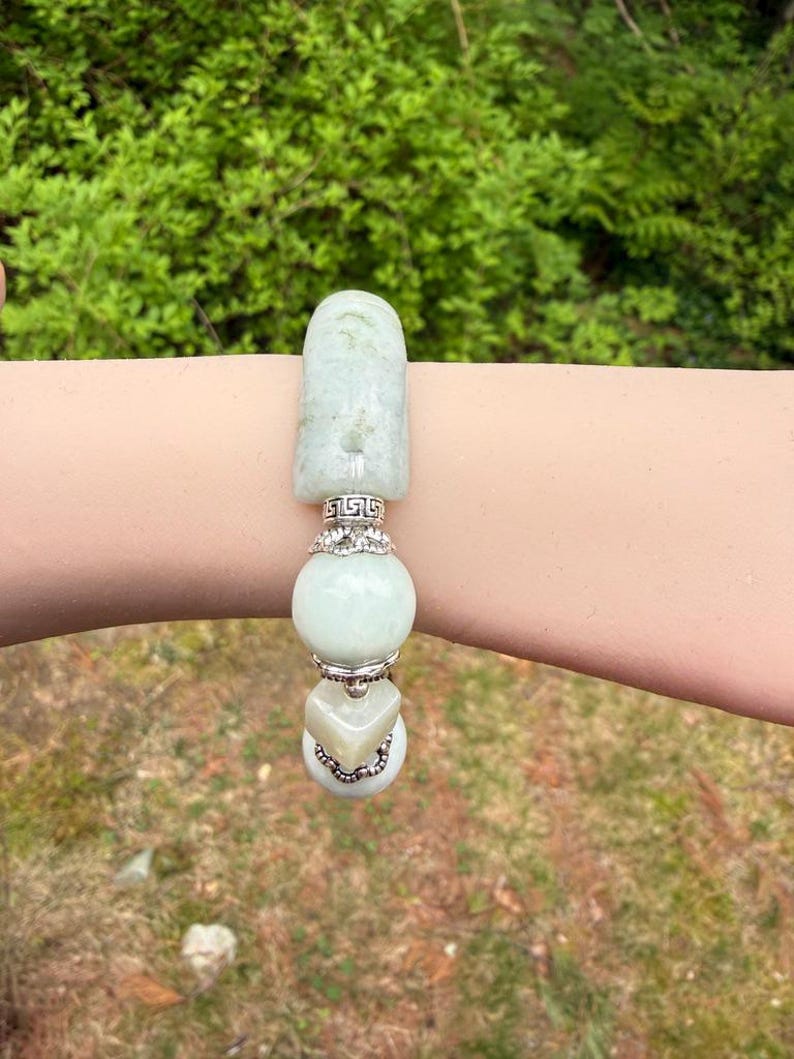 K&ouml;nnte beinhalten: Ein hellgr&uuml;nes Jade-Armband mit silbernen Akzenten. Das Armband hat ein rechteckiges Oberteil, eine runde Perle und eine herzf&ouml;rmige Perle. Das Armband wird an einem Arm vor dem Hintergrund gr&uuml;ner Bl&auml;tter gezeigt.