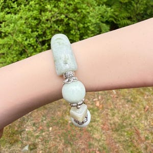 K&ouml;nnte beinhalten: Ein hellgr&uuml;nes Jade-Armband mit silbernen Akzenten. Das Armband besteht aus einem rechteckigen Jade-Element, einer gro&szlig;en runden Perle und einer quadratischen Perle. Silberne Akzente umfassen dekorative B&auml;nder und Verbinder. Das Armband wird vor einem nat&uuml;rlichen gr&uuml;nen Hintergrund gezeigt.