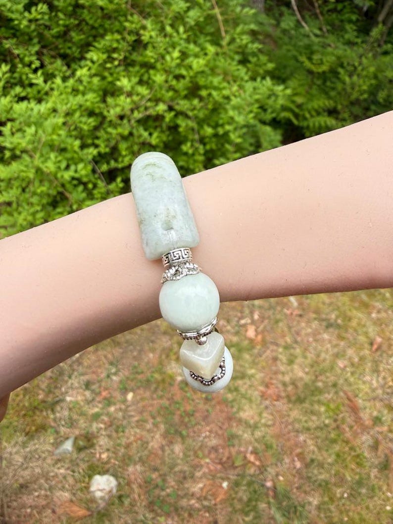 K&ouml;nnte beinhalten: Ein hellgr&uuml;nes Jade-Armband mit silbernen Akzenten. Das Armband besteht aus einem rechteckigen Jade-Element, einer gro&szlig;en runden Perle und einer quadratischen Perle. Silberne Akzente umfassen dekorative B&auml;nder und Verbinder. Das Armband wird vor einem nat&uuml;rlichen gr&uuml;nen Hintergrund gezeigt.