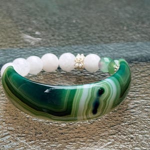 Puede incluir: Una pulsera que presenta un gran segmento curvo de ágata pulida con bandas verdes y blancas, conectado a una hilera de cuentas lisas de color rosa pálido, un pequeño espaciador dorado con diminutas perlas blancas y dos cuentas verdes translúcidas.