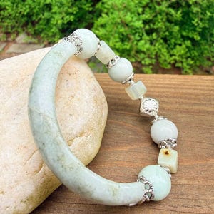 K&ouml;nnte beinhalten: Ein hellgr&uuml;nes Jade-Armband mit silberfarbenen Akzenten. Das Armband besteht aus einem gebogenen Jadeteil und einer Reihe von runden und quadratischen Jadeperlen. Die Perlen sind durch silberfarbene Filigranarbeiten verbunden. Das Armband wird auf einer Holzoberfl&auml;che pr&auml;sentiert.