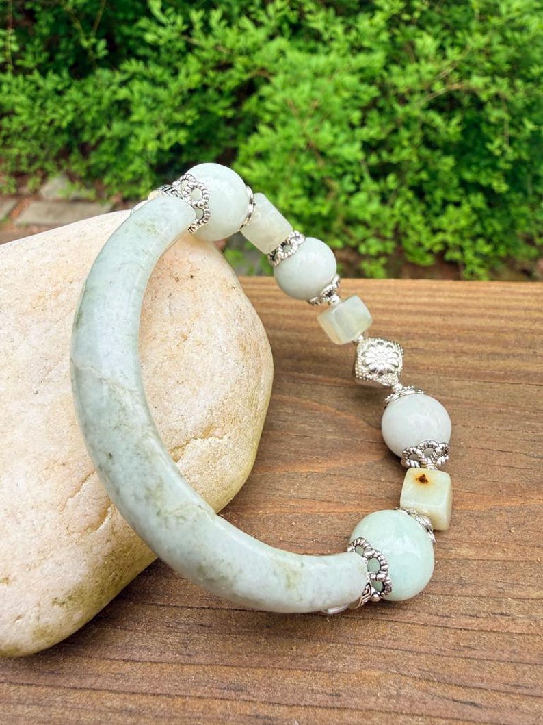 K&ouml;nnte beinhalten: Ein hellgr&uuml;nes Jade-Armband mit silberfarbenen Akzenten. Das Armband besteht aus einem gebogenen Jadeteil und einer Reihe von runden und quadratischen Jadeperlen. Die Perlen sind durch silberfarbene Filigranarbeiten verbunden. Das Armband wird auf einer Holzoberfl&auml;che pr&auml;sentiert.