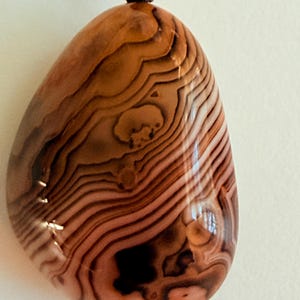 May include: A polished, teardrop-shaped pendant with a unique pattern of brown and tan wavy lines. The pendant is attached to a dark brown cord and a small, round, red bead.