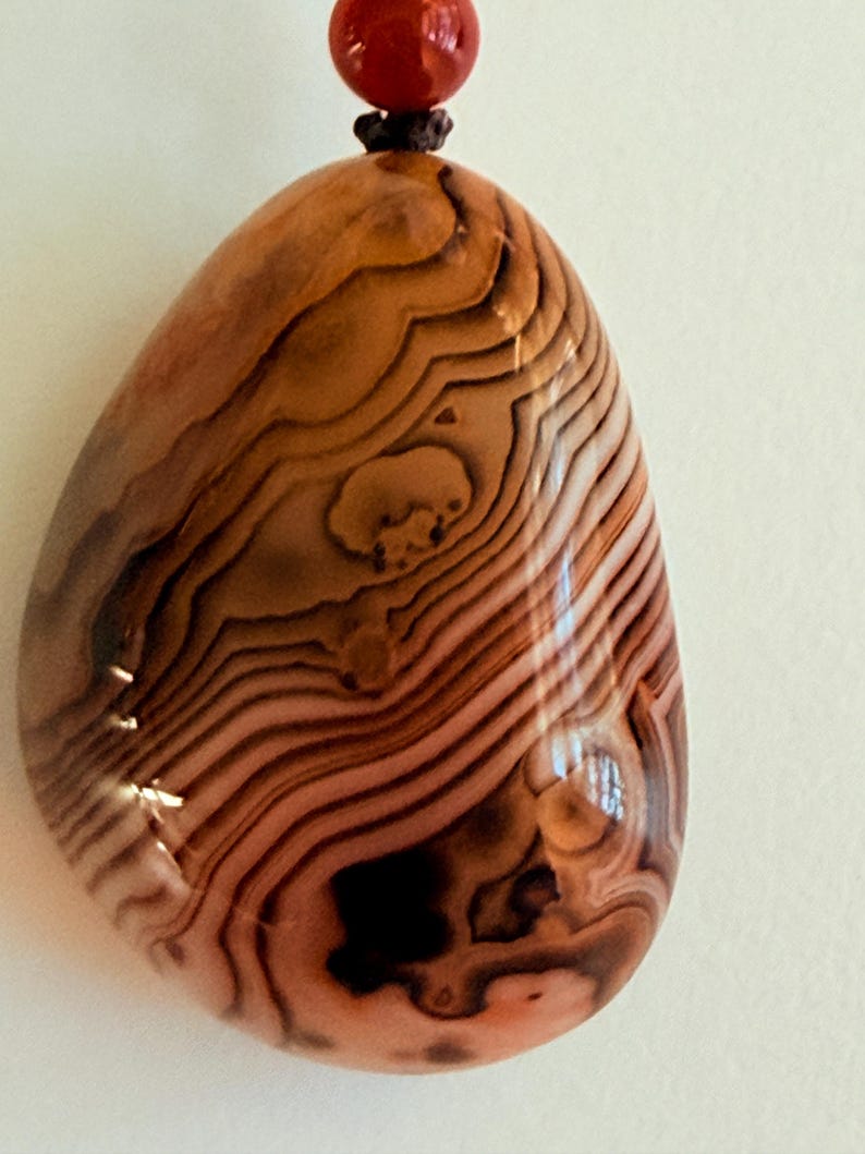 May include: A polished, teardrop-shaped pendant with a unique pattern of brown and tan wavy lines. The pendant is attached to a dark brown cord and a small, round, red bead.