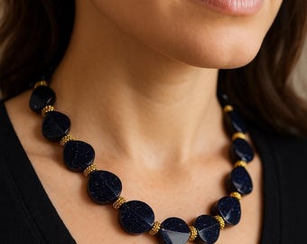 Puede incluir: Un collar de cuentas azul oscuro con detalles dorados. El collar presenta cuentas grandes, redondas y ovaladas con un aspecto moteado. Cuentas doradas separan las cuentas más grandes. El collar se usa sobre una blusa negra.