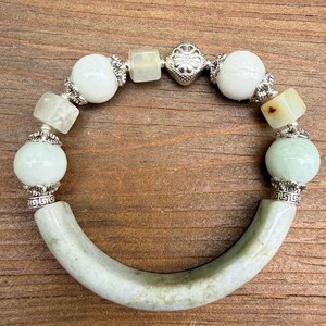 K&ouml;nnte beinhalten: Armband aus Jade und Silber mit einem gebogenen Jadest&uuml;ck und einer Reihe wei&szlig;er und gr&uuml;ner Perlen. Das Armband hat silberne Akzente und wird auf einer Holzoberfl&auml;che gezeigt.