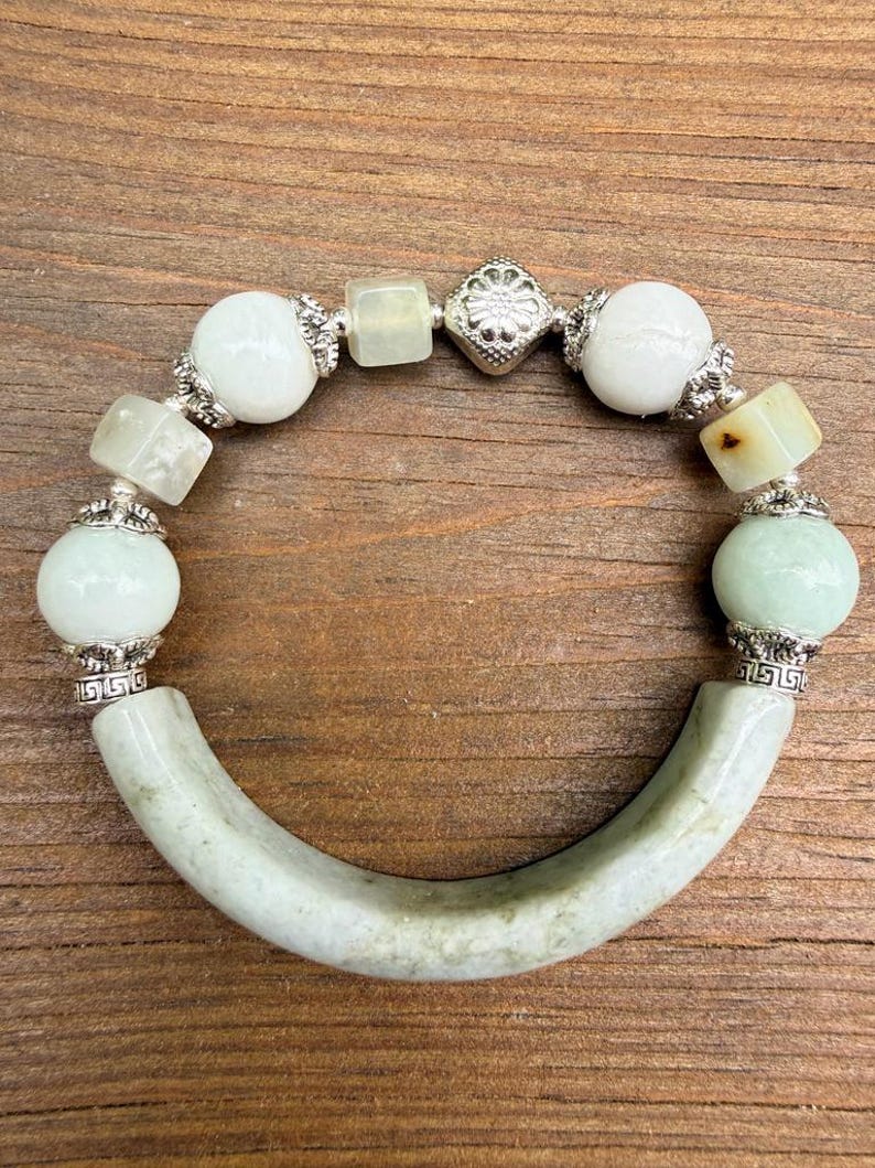 K&ouml;nnte beinhalten: Armband aus Jade und Silber mit einem gebogenen Jadest&uuml;ck und einer Reihe wei&szlig;er und gr&uuml;ner Perlen. Das Armband hat silberne Akzente und wird auf einer Holzoberfl&auml;che gezeigt.