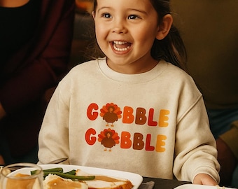 Op de afbeelding: Een kind draagt een beige sweatshirt met de woorden "GOBBLE GOBBLE" in oranje en rode letters. Het kind lacht aan een Thanksgiving-dinnertafel met eten en decoraties.