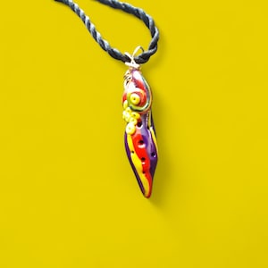 May include: A colorful pendant necklace with a unique, abstract design. The pendant features a combination of red, yellow, purple, and green hues, with a black cord. The pendant is suspended from a twisted black cord, creating a striking contrast against the yellow background.