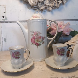May include: A white porcelain tea set with a floral rose design. The set includes a coffee pot, two cups, and saucers, all with gold trim. The background features a white wooden surface and a decorative wreath.
