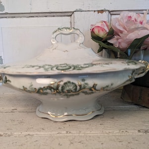 May include: White porcelain serving dish with a lid and handles. The dish features green and gold floral detailing. The lid has a handle. The dish is on a white surface.