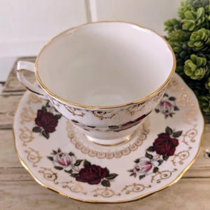 Puede incluir: Un juego de taza y platillo blancos con borde dorado y estampado de rosas. Las rosas son de color rojo intenso y rosa, con hojas verdes. El platillo tiene un borde festoneado y ribete dorado.