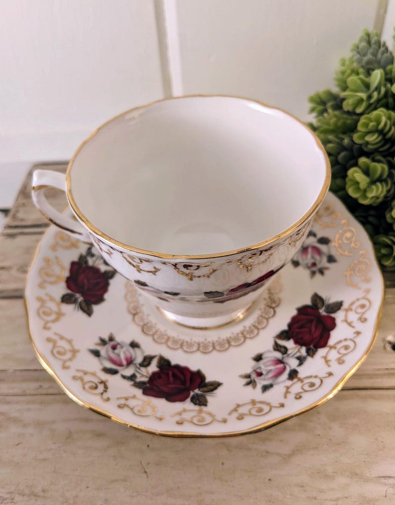 Puede incluir: Un juego de taza y platillo blancos con borde dorado y estampado de rosas. Las rosas son de color rojo intenso y rosa, con hojas verdes. El platillo tiene un borde festoneado y ribete dorado.