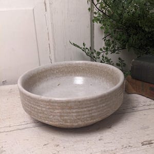 May include: A round, off-white ceramic bowl with a textured, horizontal striped pattern on the exterior. The interior is a lighter shade of white. The bowl sits on a distressed, white-painted wooden surface.