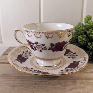 Puede incluir: Juego de taza y platillo blancos con un estampado floral de rosas en burdeos y rosa. La taza y el platillo tienen ribetes y detalles dorados. La taza est&aacute; sobre una superficie de madera.