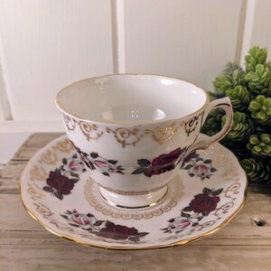 Puede incluir: Juego de taza y platillo blancos con un patr&oacute;n floral de rosas burdeos y rosas, acentuado con ribetes dorados. La taza tiene un asa delicada y se asienta sobre un platillo a juego, creando una est&eacute;tica vintage elegante.
