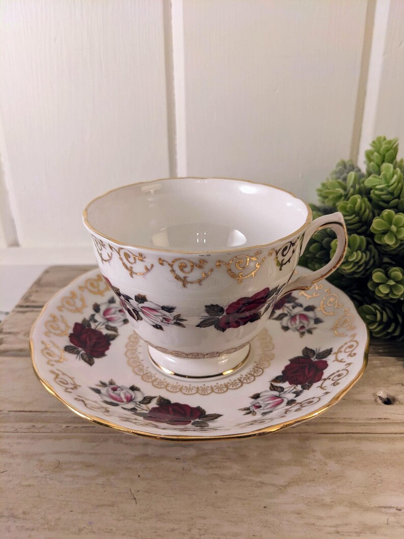 Puede incluir: Juego de taza y platillo blancos con un patr&oacute;n floral de rosas burdeos y rosas, acentuado con ribetes dorados. La taza tiene un asa delicada y se asienta sobre un platillo a juego, creando una est&eacute;tica vintage elegante.
