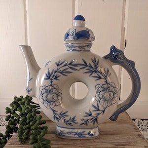 May include: White and blue ceramic teapot with a unique donut-shaped design. The teapot features blue floral and leaf patterns, a curved handle, and a lid with a blue knob. The spout is on the left side. A decorative piece for the kitchen.
