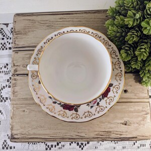 Puede incluir: Un juego de taza y platillo blanco con borde dorado y detalles florales. El platillo presenta rosas rojas y adornos dorados. La taza est&aacute; centrada en el platillo, creando un aspecto cl&aacute;sico y elegante. El juego se coloca sobre una superficie de madera r&uacute;stica.