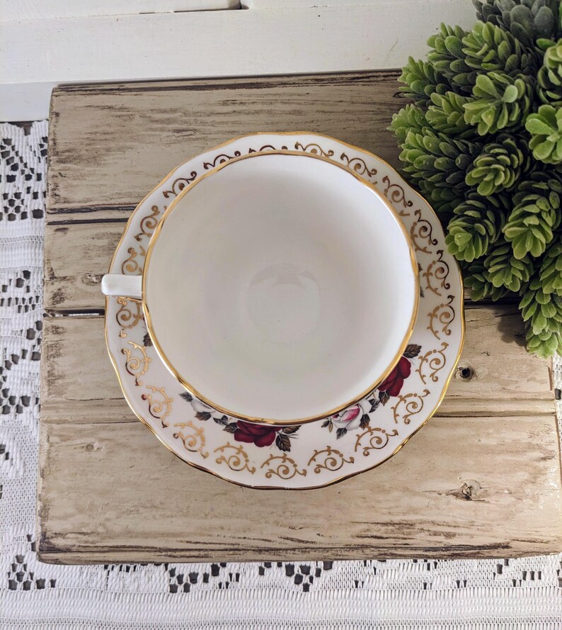 Puede incluir: Un juego de taza y platillo blanco con borde dorado y detalles florales. El platillo presenta rosas rojas y adornos dorados. La taza est&aacute; centrada en el platillo, creando un aspecto cl&aacute;sico y elegante. El juego se coloca sobre una superficie de madera r&uacute;stica.