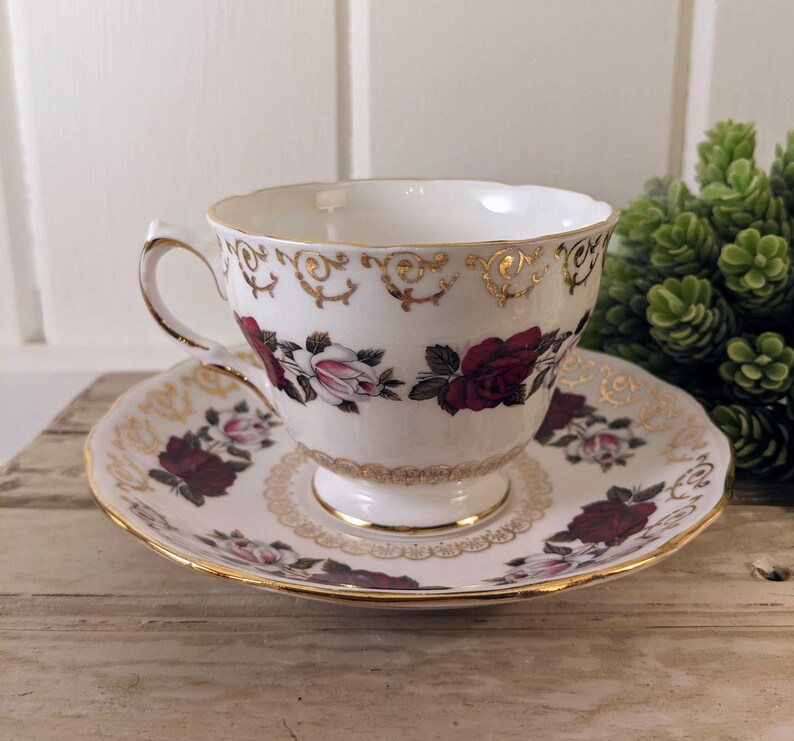 Puede incluir: Juego de taza y platillo blancos con un patr&oacute;n floral de rosas burdeos y rosas. La taza y el platillo tienen detalles y bordes dorados. Colocado sobre una superficie de madera.
