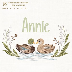 Könnte beinhalten: Stickdesign für Maschinen mit zwei sich zugewandten Enten und dem Namen "Annie" darüber. Das Design enthält florale Elemente und ist in den Größen 10 cm, 13 cm, 15 cm, 18 cm und 20 cm erhältlich.