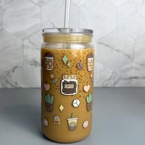 May include: A clear glass tumbler with a bamboo lid and a clear straw, filled with a coffee-colored beverage. The tumbler is decorated with coffee-themed stickers, including coffee cups, coffee beans, and flowers. The tumbler is approximately 6 inches tall.