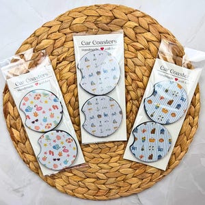May include: Six circular car coasters packaged in three clear plastic sleeves, each labeled Car Coasters. The coasters feature various summer-themed patterns including beach chairs, flamingos, sunglasses, and nautical icons on white or blue striped backgrounds.