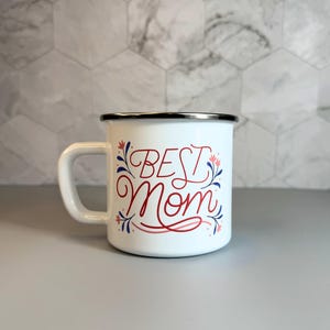 May include: White enamel mug with a black rim and a white handle. The mug features the words "BEST Mom" in red script, accented with blue and pink floral designs. A perfect gift for Mother's Day.