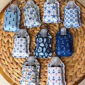 May include: A collection of small, refillable hand sanitizer bottles in decorative fabric holders with keychains. The holders feature various nautical patterns, including anchors, seashells, lighthouses, and starfish, in shades of blue and white.