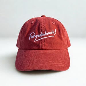 Puede incluir: Gorra de béisbol de pana roja con el texto bordado "Fuhgedaboutit!" en escritura blanca. La gorra tiene una visera curva y un botón en la parte superior. El sombrero es de color rojo intenso.