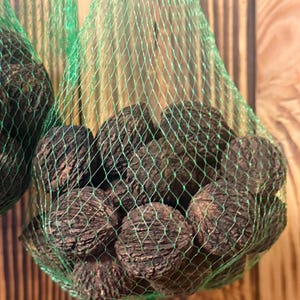 以下が含まれることがあります： 緑色のメッシュバッグに入った濃い茶色のクルミのクローズアップ。クルミはざらざらとした質感の表面をしています。緑色のメッシュバッグは木製の背景に吊り下げられています。
