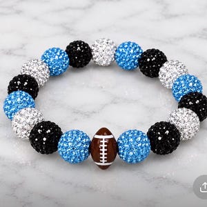 May include: A stretch bracelet featuring alternating round, textured beads in black, white, and bright blue. The beads have a sparkling, glittery finish. A single brown football-shaped bead with white lace detailing is centered in the design.