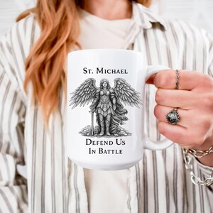 Peut inclure: Mug en céramique blanche avec une illustration détaillée de Saint Michel, un ange avec des ailes, tenant une épée. Le texte "St. Michael" et "Defend Us In Battle" sont imprimés sur le mug. La tasse a une grande anse.