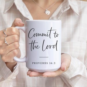Puede incluir: Taza de cerámica blanca con el texto "Commit to the Lord" en escritura negra, con "Proverbios 16:3" debajo. La taza es sostenida por una persona que lleva una camisa de color claro.