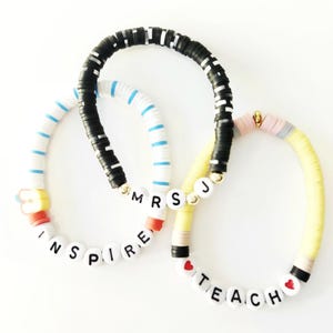 May include: Three colourful beaded bracelets. One black and white bracelet, one white and blue bracelet with the word "INSPIRE", and one yellow bracelet with the words "MRS TEACH". The bracelets have gold accents and red heart beads.
