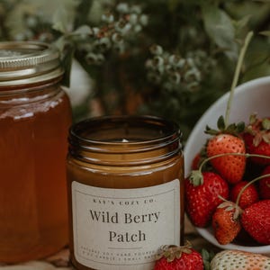 Pode incluir: Uma vela num frasco de vidro castanho com um rótulo creme que diz Kay's Cozy Co. Wild Berry Patch Natural Soy Hand Poured 8 oz Candle Small Batch. A vela está ao lado de um frasco de mel e de uma taça branca com morangos frescos.