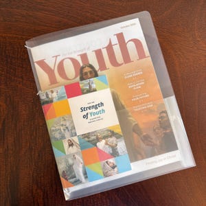 May include: A clear plastic document sleeve containing the October 2026 issue of the For the Strength of Youth magazine and a For the Strength of Youth guide for making choices booklet. The magazine features religious imagery and text on the cover.