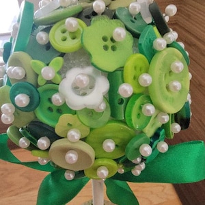 May include: A decorative topiary made of various shades of green buttons and faux pearls. The buttons are arranged in a spherical shape, with a white stem and a green ribbon bow at the base. The topiary sits on a bed of green moss.