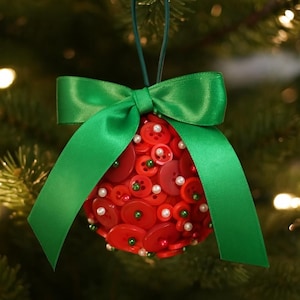 May include: A festive Christmas ornament, spherical in shape, is adorned with numerous red buttons of varying sizes. Small white and green beads are interspersed among the buttons. A vibrant green satin ribbon forms a large bow at the top, completing the holiday decoration.