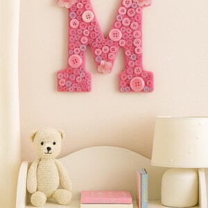 May include: A pink letter "M" made of buttons hangs on a wall. The letter is decorated with various shades of pink buttons and small flower accents. A shelf below holds a teddy bear, books, and a lamp.