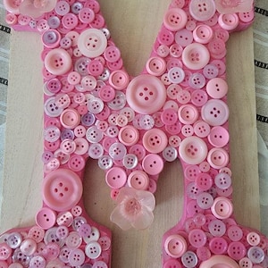 May include: Pink wooden letter "M" decorated with various sizes of pink and white buttons. The letter is adorned with several decorative pink flower accents. A unique and charming decorative piece.