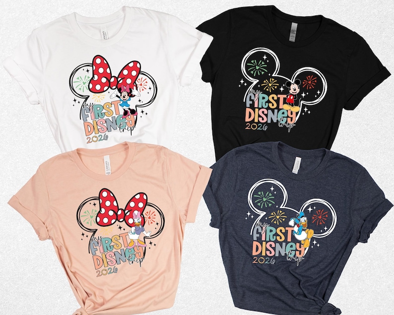 May include: Four t-shirts in white, black, peach, and navy blue. Each shirt features a Mickey Mouse head outline with fireworks and the text "First Disney 2026." Minnie Mouse, Mickey Mouse, and Donald Duck are featured in the designs.