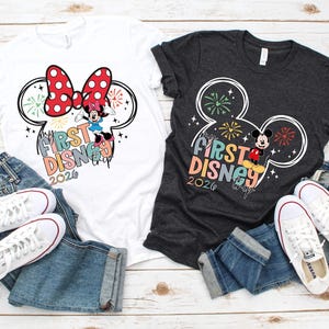 May include: Two t-shirts, one white and one charcoal gray, featuring a Mickey Mouse ear design with fireworks and the text "My First Disney Trip 2020." The shirts are paired with blue jeans and white sneakers.